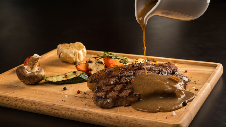 Gravy is poured onto a grilled steak on a wooden board
