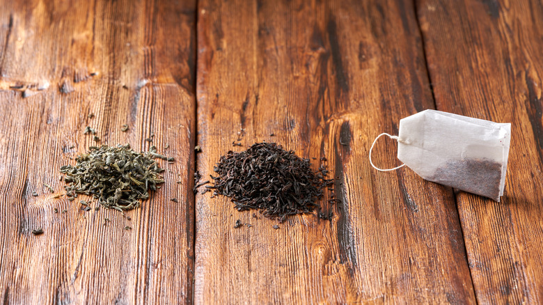 A pile of loose green tea, a pile of loose black tea, and a tea bag sit on a wooden table.