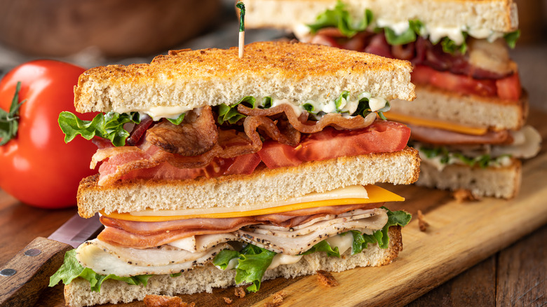 Two halves of a club sandwich on a cutting board with a tomato in the background