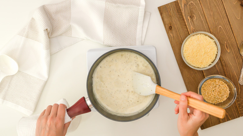 A chef mixing fresh Alfredo sauce with bowls of shredded parmesan cheese and herbs nearby.