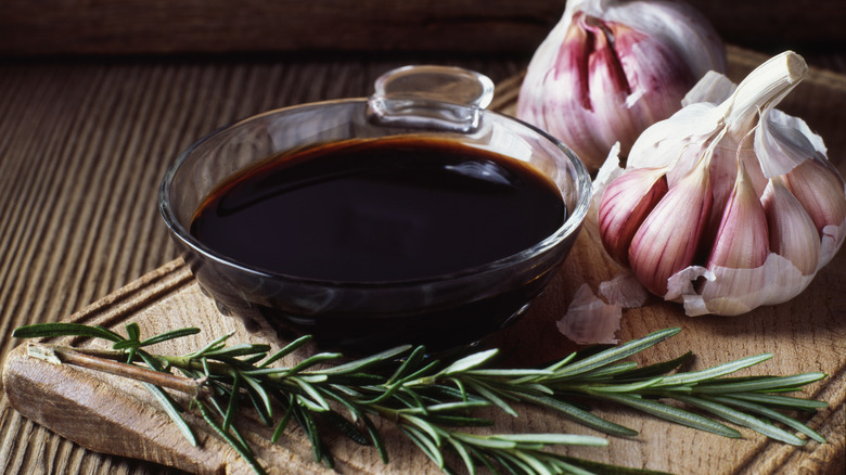 Glass bowl of balsamic vinegar on a wooden cutting board with two heads of garlic and a sprig of fresh rosemary