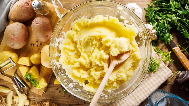 A bowl of mashed potatoes with potatoes and herbs next to it