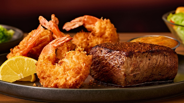 Steak on a plate with fried shrimp, lemon wedges, and dipping sauce; salad, and broccoli are in the background.