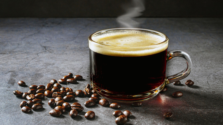 Close up of a glass mug of black coffee next to scattered coffee beans