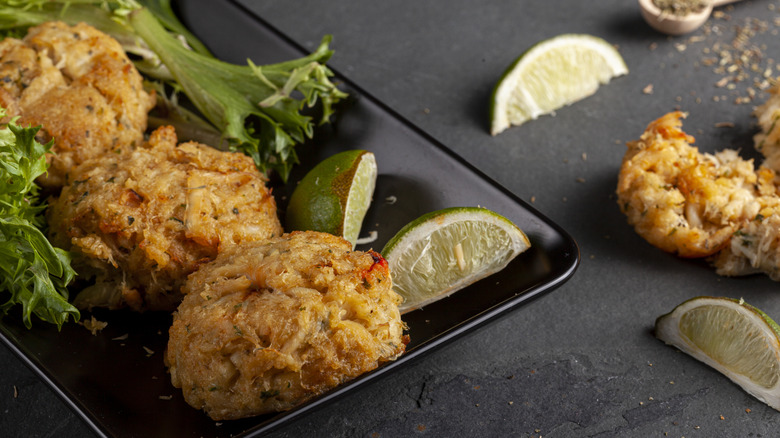 A plate of crab cakes with lime wedges sits on a black table