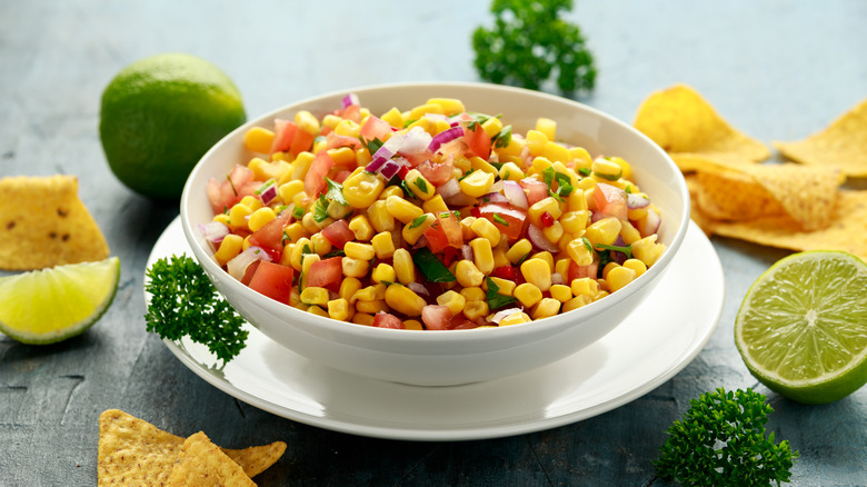 White bowl of corn salsa with lime wedges and halves, fresh parsley, and tortillas on the side