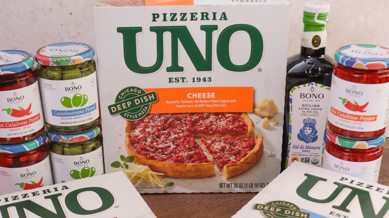 Box of Pizzeria Uno Cheese frozen pizza sits on a table surrounded by other jarred peppers and olive oil