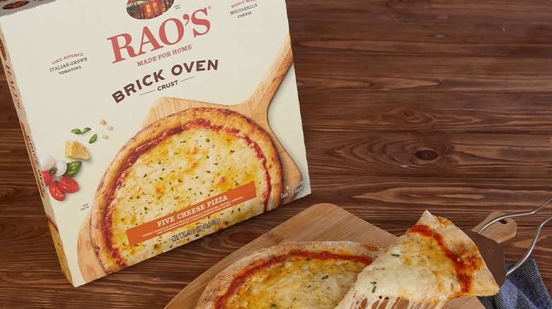 A box of Rao's Brick Oven Five Cheese Pizza sits upright on a table next to a cooked pizza with one slice being pulled out