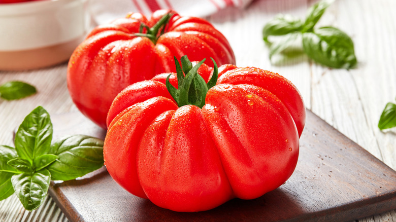 Two large, bright red heirloom tomatoes on a wooden cutting board, surrounded by basil leaves