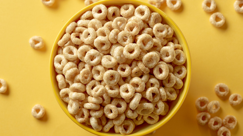 A yellow bowl filled with Cheerios sits on a yellow surface, with individual Cheerios scattered around the bowl