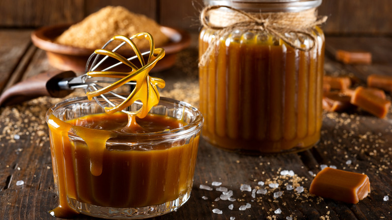 Two jars of caramel sauce, one with a whisk dripping with the sauce, on a table with salt flakes and caramel squares