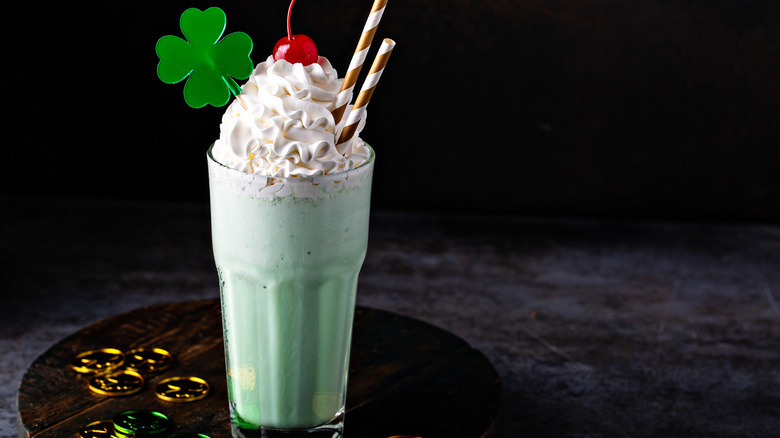 A green shake with a shamrock decoration has whipped cream and a cherry on top while sitting next to golden and green cins