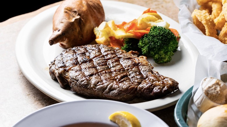 Steak on a plate next to a baked potato, broccoli, and assorted veggies from Montana Mike's Steakhouse.