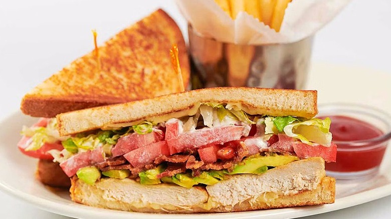 A Cheesecake Factory Grilled Chicken and Avocado Club sandwich sits on a plate with fries and ketchup