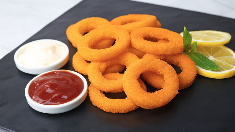 Pile of onion rings on a table next to mayo, ketchup, and lemon slices