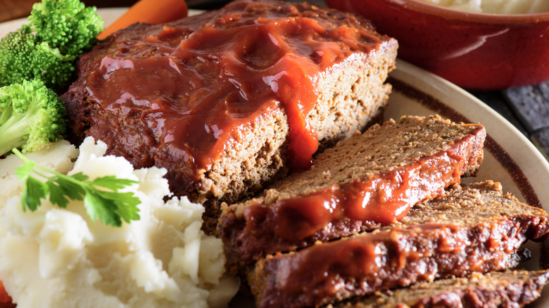 A meatloaf, half whole and half in slices, sits on a plate alongside mashed potatoes, broccoli, and carrots