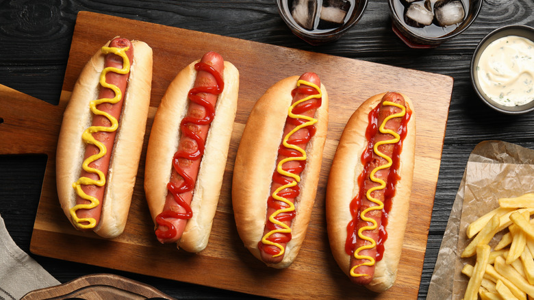 hot dogs topped with ketchup and mustard sit on a wooden board next to glasses of soda, fries, and a bowl of dipping sauce
