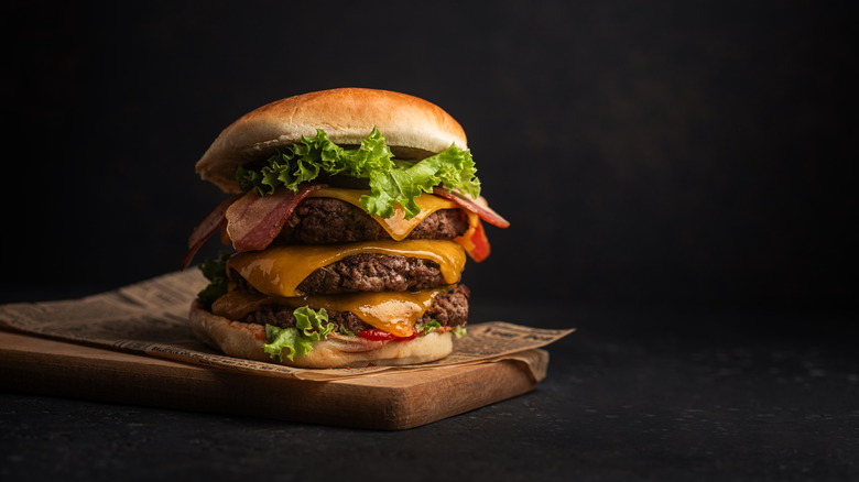 A huge three-patty burger sits on a wooden cutting board, with a dark background