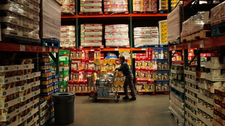A Costco shopper with short hair and glasses pushes a shopping cart down an aisle