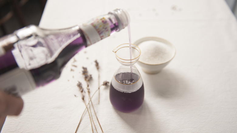 Someone pours lavender syrup into a glass jug, with fresh lavender and a small bowl of sugar beside it