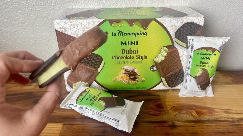 Hand holding La Menorquina Dubai Chocolate-Style Mini Ice Cream Sandwich with packaging and wrapped ones in the background