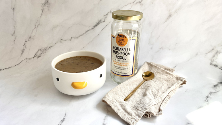 A bowl of Zoup! Portabella Mushroom Bisque next to the packaging and a golden spoon on a light colored cloth napkin.