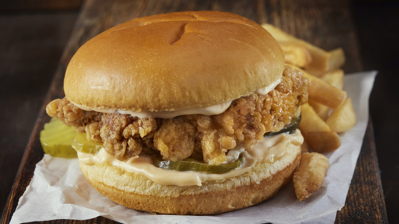 A fast food crispy chicken sandwich with pickles and sauce, sitting on white parchment paper