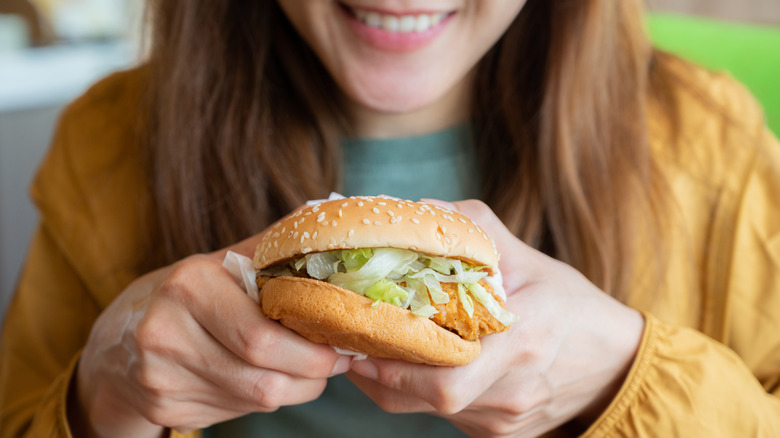 A woman smiles while holding a fast food chicken sandwich with shredded lettuce