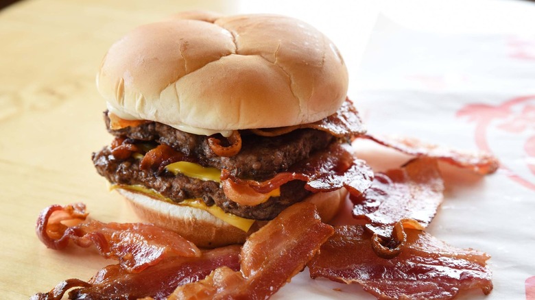 A Baconator sits on its wrapper with bacon in front of it
