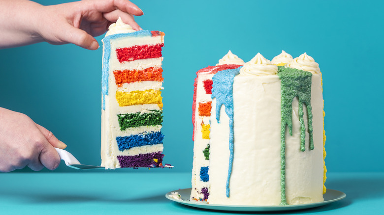 Hands remove a tall wedge of rainbow layer cake from the rest of the cake using a serving utensil