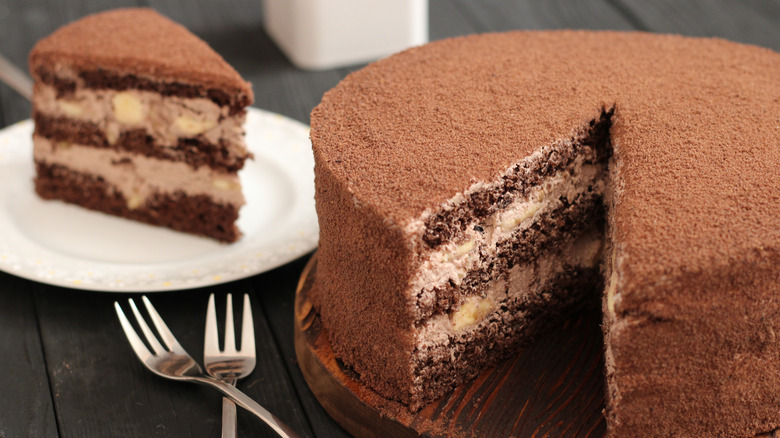 A layered chocolate cake sits on a round, wooden serving dish, with two forks lying adjacent and a slice of the cake on a white plate in the background