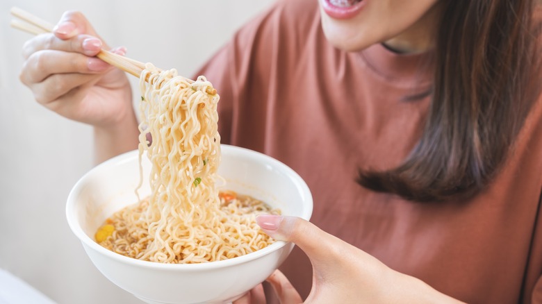 A woman smiles while holding a white bowl full of instant ramen