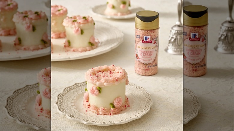 A shaker bottle of Bridgerton Strawberries & Cream Finishing Sugar sits next to plates of small, ornate, pink-and-white frosted cakes