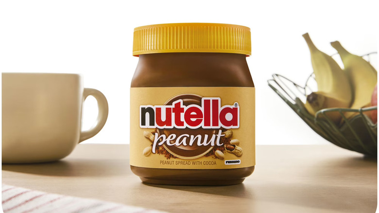 A jar of Nutella Peanut sits next to a coffee cub and a bowl of fruit.
