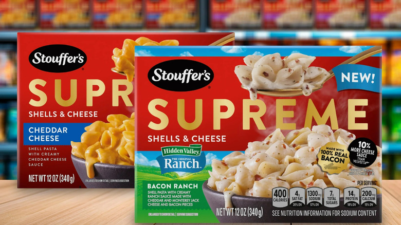 A box of Stouffer's Supreme Bacon Ranch Shells & Cheese and Original Shells & Cheese sit in front of a supermarket freezer case