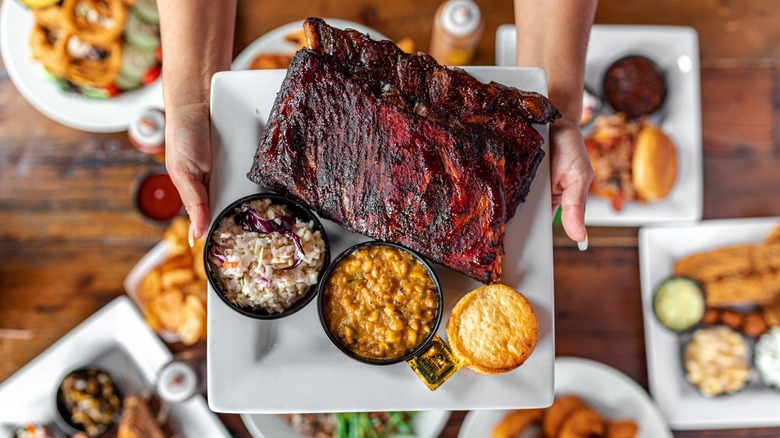 Hands holding plate full of Smokin' Dave's BBQ with sides over collection of other dishes