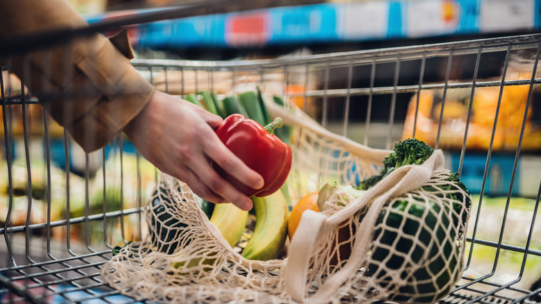 A hand sets a red pepper into a reusable net bag of produce inside a grocery store cart