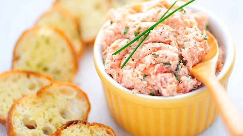 A salmon spread with herb garnishes sits in a yellow ramekin with a wooden spoon, surrounded by slices of baguette