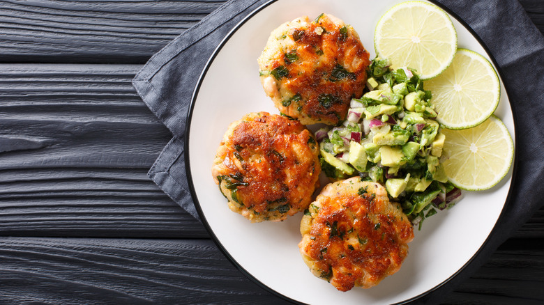 Three salmon cakes on a white plate, garnished with lemon slices and an avocado salsa
