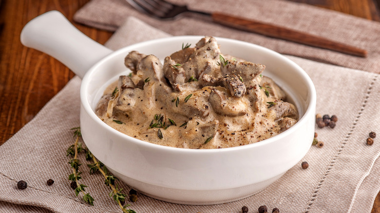 Beef stroganoff sits in a white tureen on a burlap napkin surrounded by fresh thyme and whole peppercorns