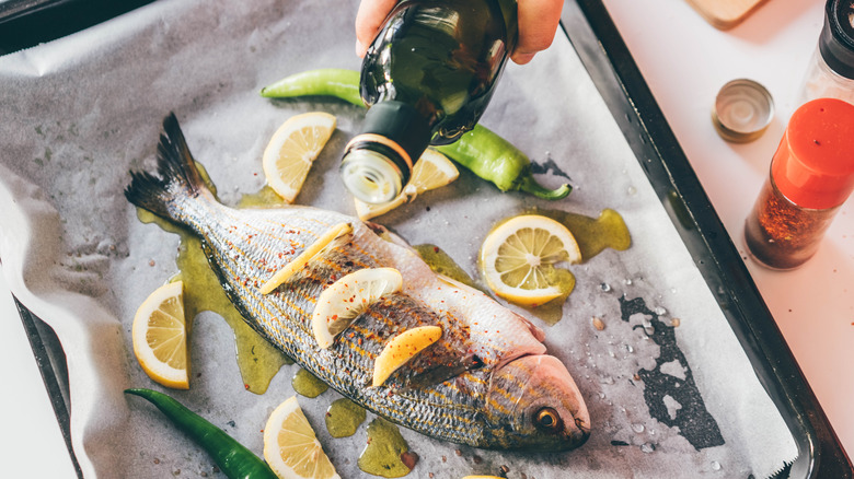 A whole fish, surrounded by accoutrements, sits on paper on a baking sheet as a hand drizzles olive oil