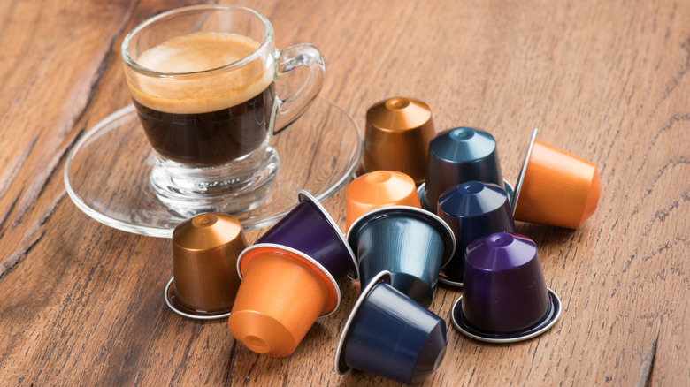 A cup of espresso sits in a glass saucer, near a group of Nespresso pods