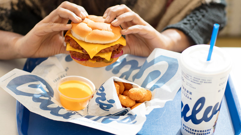 Hands hold a Culver's ButterBurger above a food tray containing cheese curds, sauce, and a drink