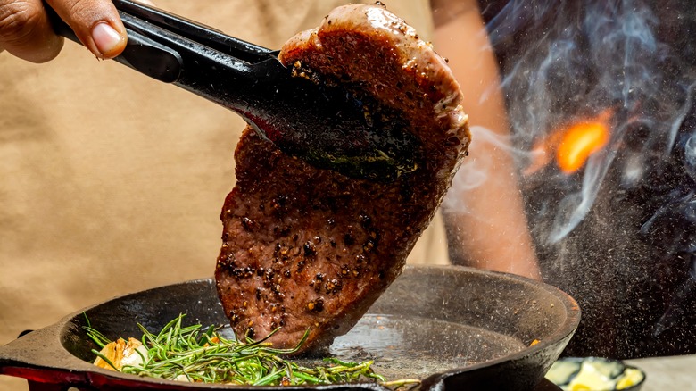 A hand flipping a steak while it is cooking, with a pair of tongs