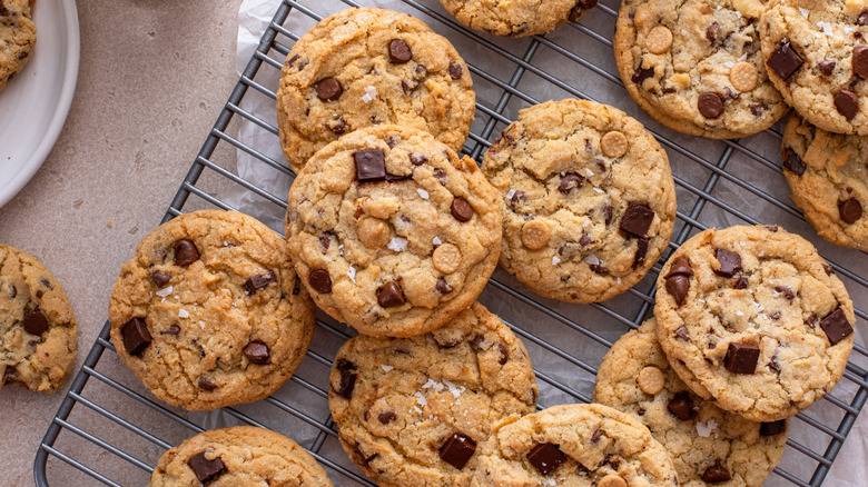 A large batch of cookies with chocolate chips, butterscotch chips, and sea salt sit on a cooling rack