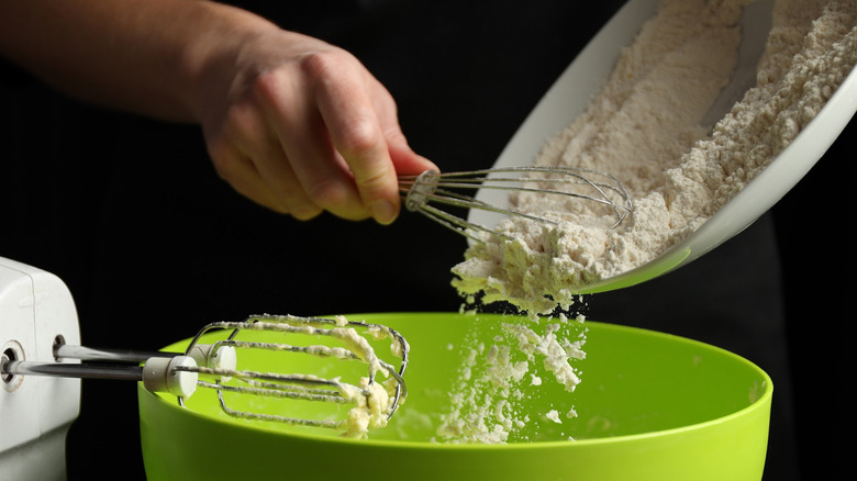 A chef holds a whisk while pouring a plate of flour into a green bowl filled with batter