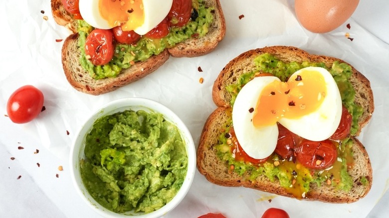 Slices of seeded hero bread with avocado mash, roasted tomatoes, jammy eggs, and chili flakes.