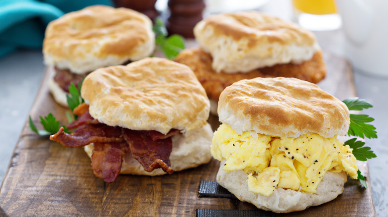 Four breakfast biscuit sandwiches with eggs, bacon, and chicken on a wooden board