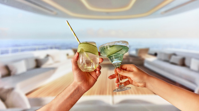 Two people clink glasses that have boozy drinks that feature cucumber on a yacht