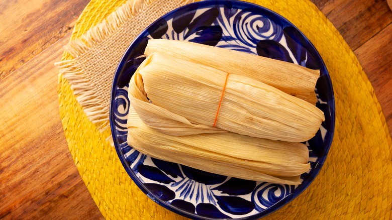 Multiple tamales wrapped in corn husks sit on a blue patterned plate on a dining table
